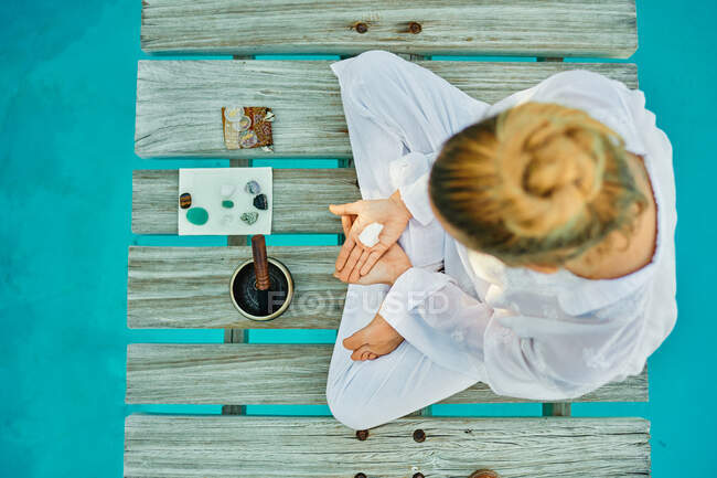 From above unrecognizable blond male hipster yogi in white clothes sitting in lotus pose meditating near tibetan singing bowl and crystals on wooden path bridge on top of a turquoise pool during spiritual retreat - foto de stock