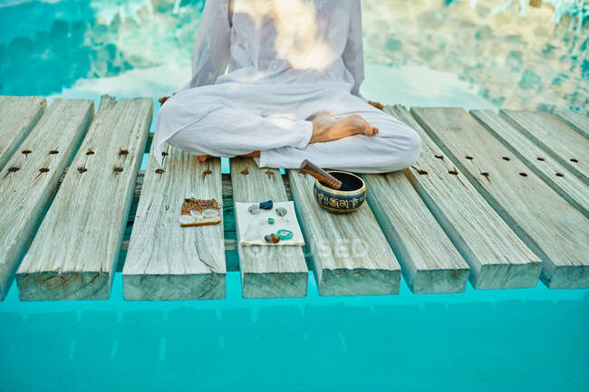 Zugeschnittene männliche Hipster-Yogi in weißen Kleidern sitzen in Lotus-Pose und meditieren mit Kristallen in der Nähe der tibetischen Klangschale auf einer hölzernen Pfadbrücke auf einem türkisfarbenen Pool während des spirituellen Retreats — Stockfoto