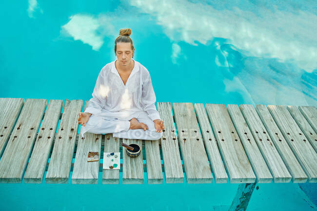 Desde arriba, el rubio hipster yogui masculino con ropa blanca sentado en una pose de loto meditando con los ojos cerrados cerca de un cuenco tibetano y cristales en un puente de madera sobre una piscina turquesa en un jardín tropical durante el retiro espiritual - foto de stock