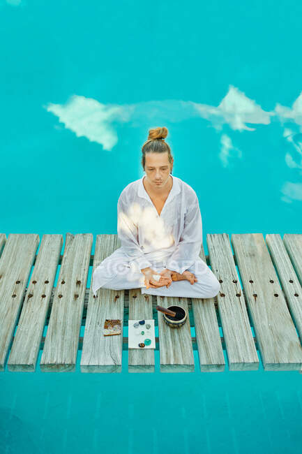D'en haut blond mâle hipster yogi en vêtements blancs assis dans la pose de lotus méditant près du bol de chant tibétain et des cristaux sur le pont de chemin en bois sur le dessus d'une piscine turquoise dans le jardin tropical pendant la retraite spirituelle — Photo de stock