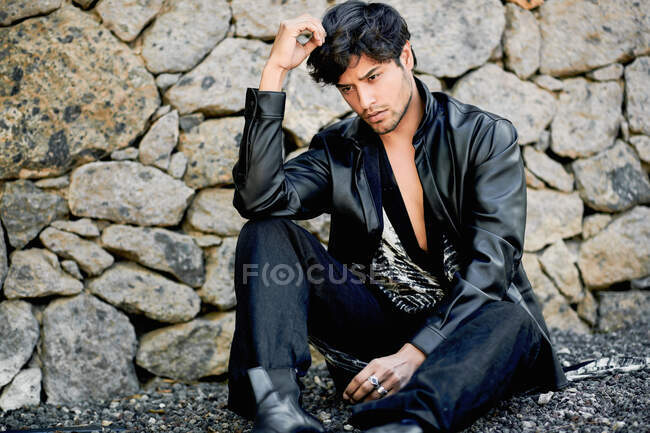 Young sensual handsome ethnic male model wearing hipster stylish summer clothes looking away while sitting pavement leaning on rustic rural stone wall — Stock Photo