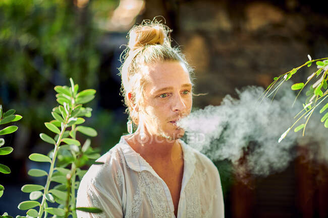 Pensativo guapo rubio macho vistiendo ropa de verano hipster mirando hacia otro lado mientras está de pie fumando cigarrillo rodeado de vegetación verde - foto de stock
