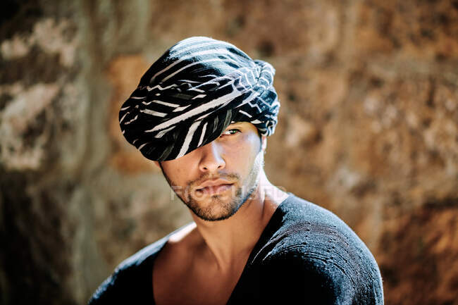 Joven sensual guapo étnico árabe masculino con turbante modelo vistiendo hipster ropa de verano elegante mirando hacia otro lado mientras está de pie en la calle - foto de stock