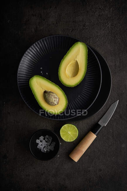 Сверху свежие половинки авокадо расположены на столе с зеленой лаймом, солью и ножом на темном фоне — стоковое фото