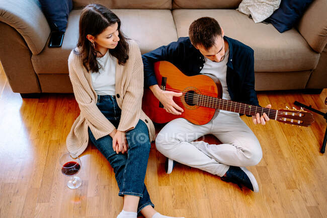 Талантливый мужчина-музыкант играет на акустической гитаре для женщины, сидящей на диване с бокалом вина и наслаждающейся песней — стоковое фото