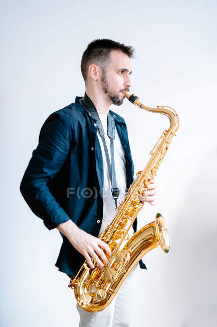 Вид збоку на творчого музиканта-чоловіка, який грає на саксофоні, стоячи на білому тлі і дивлячись в сторону — стокове фото