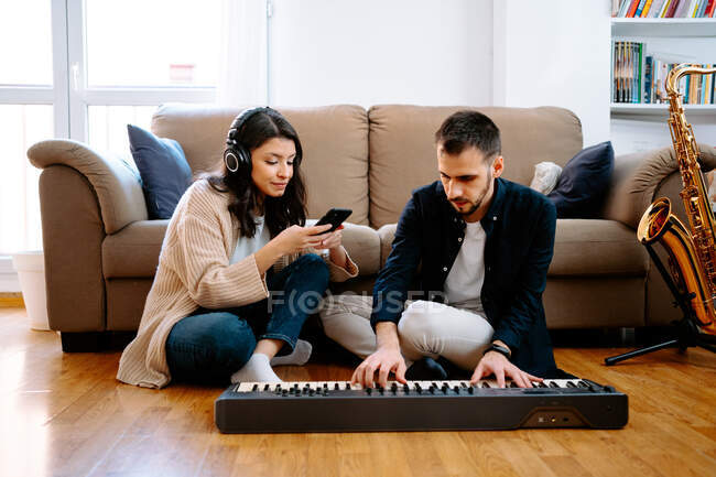 Couple of musicians sitting on floor and composing music while playing synthesizer and using smartphone — Stock Photo