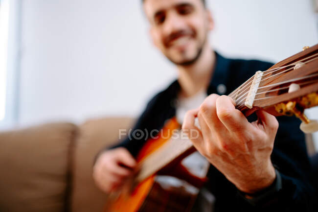 Низкий угол улыбки мужчины-музыканта, играющего на акустической гитаре, сидя дома на диване и репетируя песню — стоковое фото