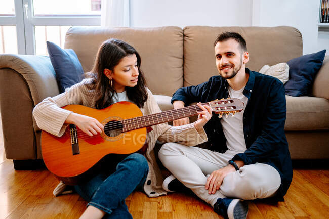 Талановита жінка-музикантка, яка грає на акустичній гітарі для чоловіка, сидить на підлозі і насолоджується піснею — стокове фото