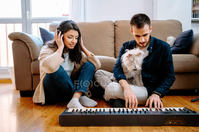 Homem tocando sintetizador e mulher ouvindo música em fones de ouvido enquanto sentado no chão em casa e gravando nova música — Fotografia de Stock