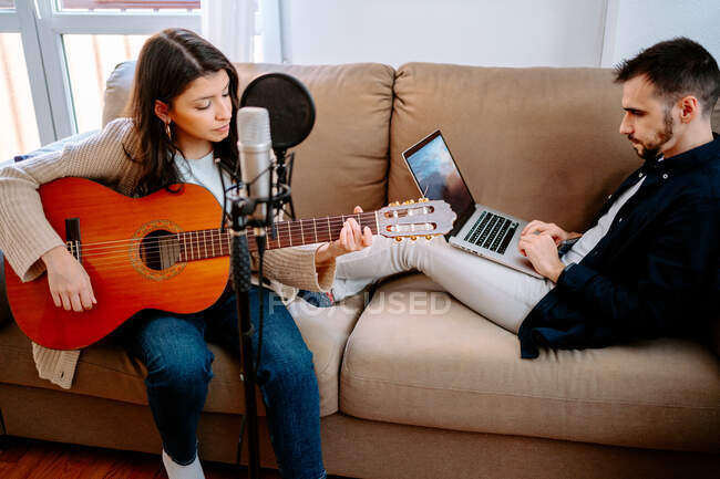 Пара музикантів сидить на дивані і записує пісню, граючи на акустичній гітарі і використовуючи ноутбук — стокове фото
