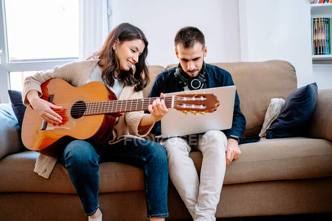 Un par de músicos sentados en el sofá y grabando canciones mientras tocan la guitarra acústica y usan el portátil - foto de stock