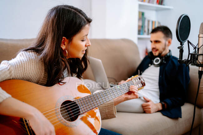 Ein paar Musiker sitzen auf dem Sofa und nehmen Songs auf, während sie akustische Gitarre spielen und Laptop benutzen — Stockfoto