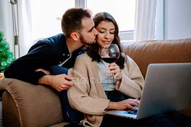 Namorado beijando conteúdo namorada sentado com vinho e laptop de navegação em casa no fim de semana — Fotografia de Stock