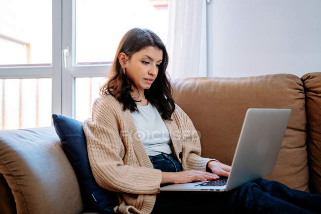Side view of focused female freelancer sitting on couch at home and working on remote project while typing on laptop — Stock Photo