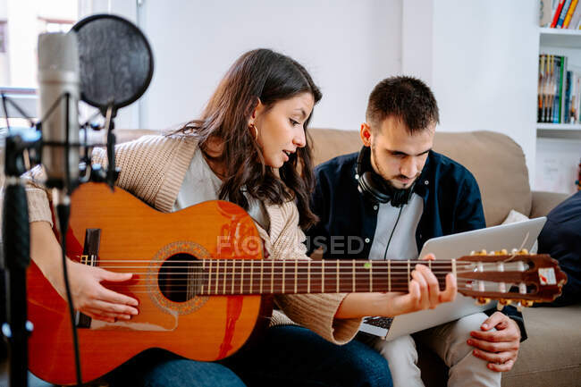Пара музикантів сидить на дивані і записує пісню, граючи на акустичній гітарі і використовуючи ноутбук — стокове фото