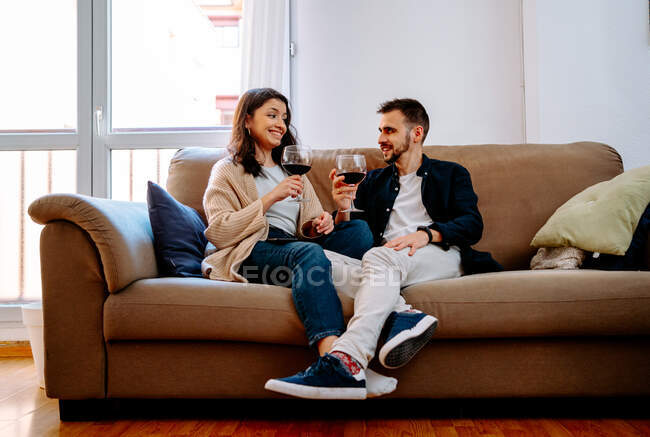Relaxed couple with red wine in glasses sitting on couch and looking at each other while spending weekend together — Stock Photo