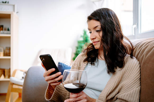 Freelancer feminino focado sentado no sofá em casa e navegando smartphone enquanto bebe vinho — Fotografia de Stock