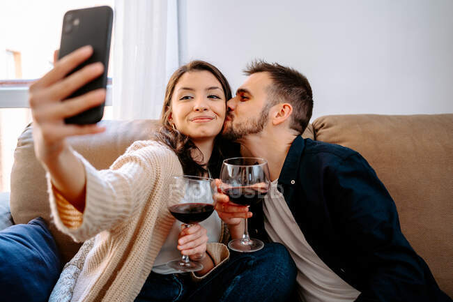 Content couple sitting on sofa with wineglasses and taking self portrait on smartphone while relaxing at weekend at home — Stock Photo