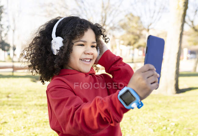 Happy ethnic child with Afro hairstyle enjoying songs in headphones while using smartphone in urban park — Stock Photo