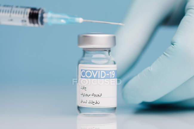 Gros plan de la culture médecin méconnaissable dans des gants seringue de remplissage avec le vaccin du coronavirus en flacon de verre sur fond bleu — Photo de stock