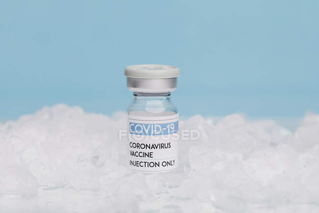 Flacon en verre avec vaccin de COVID 19 placé sur glace au congélateur sur fond bleu — Photo de stock