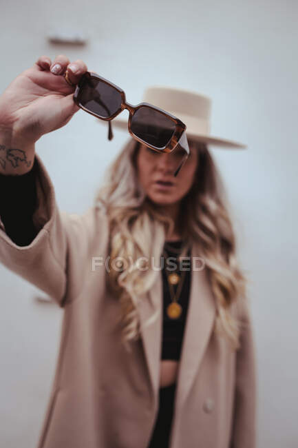 Influenciadora femenina borrosa irreconocible con cabello largo y rubio y con un atuendo elegante con sombrero y gafas de sol de pie con brazo extendido - foto de stock