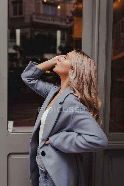 Side view of female with long blond hair and in stylish outfit standing near building in city with eyes closed — Stock Photo