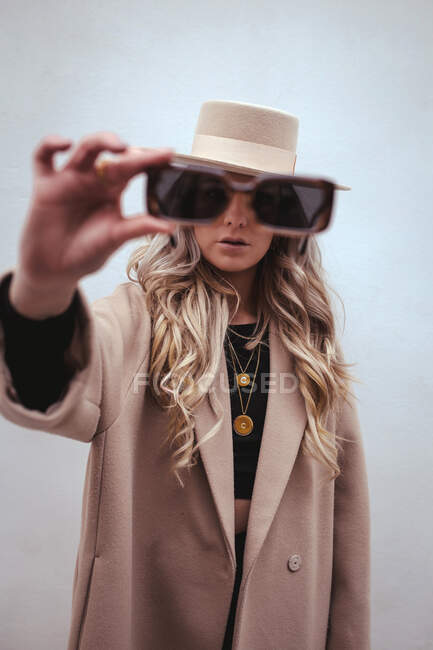 Influenciadora feminina irreconhecível com cabelo loiro longo e com roupa elegante com chapéu e óculos de sol em pé com braço estendido — Fotografia de Stock