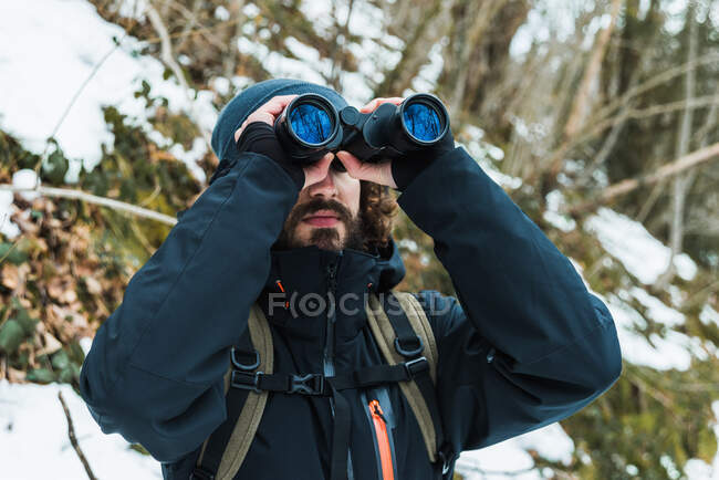 Caminhante masculino em roupas quentes em pé em bosques de inverno nevado e olhando em binóculos — Fotografia de Stock