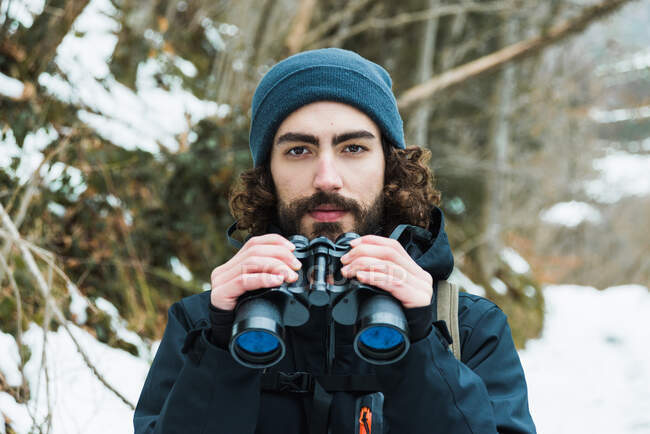 Caminhante masculino em roupas quentes em pé em bosques de inverno nevado e olhando para câmera segurando binóculos — Fotografia de Stock