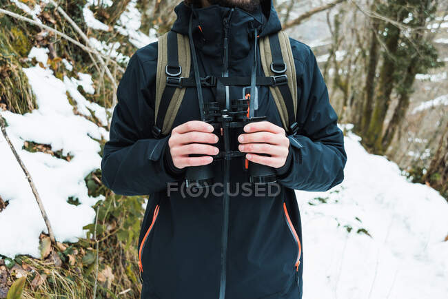 Gestutzter, bis zur Unkenntlichkeit verkleideter männlicher Wanderer in warmer Kleidung steht mit Fernglas im verschneiten Winterwald — Stockfoto