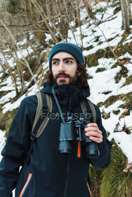 Randonneur masculin en vêtements chauds debout dans les bois enneigés d'hiver et regardant loin tenant des jumelles — Photo de stock