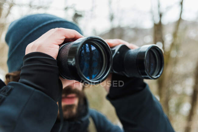 Escursionista maschio in abiti caldi in piedi in boschi invernali innevati e guardando in binocolo — Foto stock