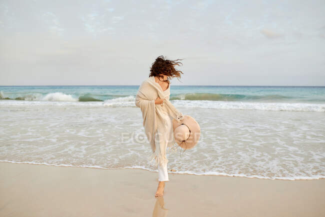 Delighted female traveler in wet clothes walking along seashore on background of rocky cliff and looking at camera — Stock Photo