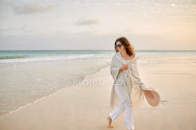 Female traveler in sunglasses and hat walking along seashore and looking away — Stock Photo
