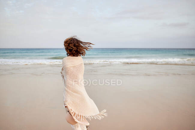 Back view female traveler holding hat and walking along seashore and looking away — Stock Photo