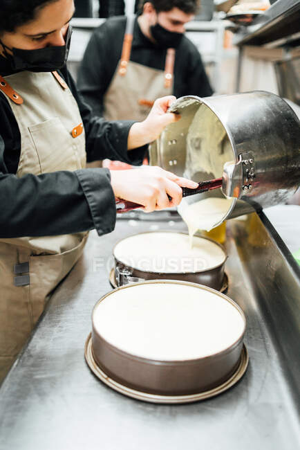 Cocinera femenina irreconocible recortada con mascarilla vertiendo crema sobre lata de pastel mientras prepara delicioso postre de pastel de queso en la cafetería - foto de stock