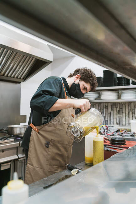 Side view of unrecognizable male bartender in apron and face mask poring fresh squeezed juice from blender into glass while working at counter in bar — Stock Photo