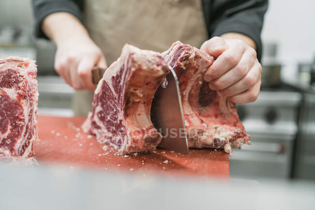 Ernte bis zur Unkenntlichkeit männlicher Metzger in Schürze schneidet Fleisch mit scharfem Messer während der Arbeit in der Küche — Stockfoto