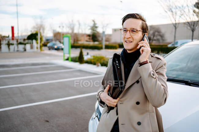 Empreendedor masculino ocupado com caso attache em pé no estacionamento perto do carro e falando no telefone celular ao discutir o projeto e olhando para longe — Fotografia de Stock