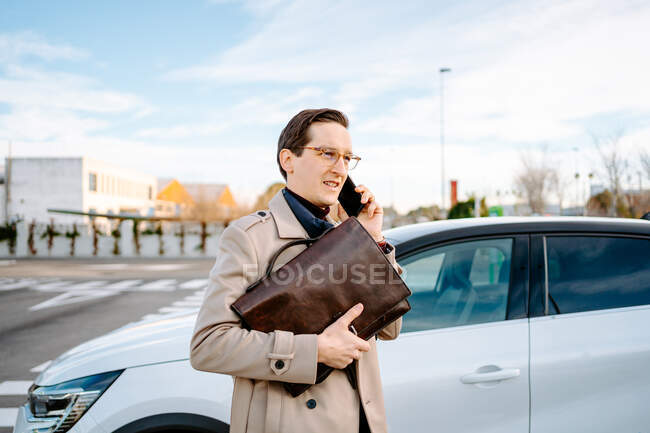 Entrepreneur masculin occupé avec attachcase debout sur le parking près de la voiture et parlant sur le téléphone mobile tout en discutant du projet et en regardant ailleurs — Photo de stock