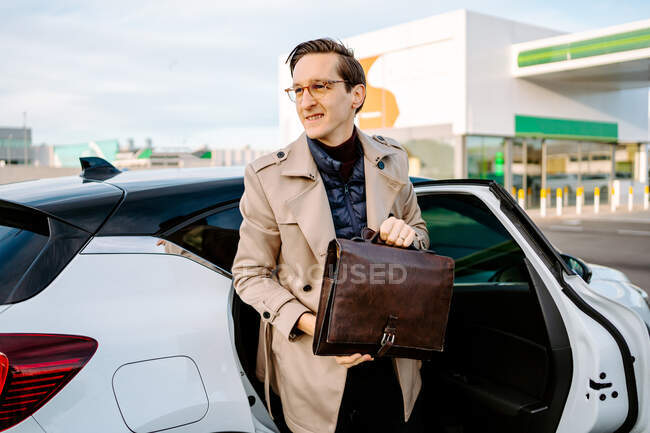 Уверенный в себе мужчина-предприниматель в пальто и с футляром атташе выходит из современного автомобиля и отворачивается — стоковое фото