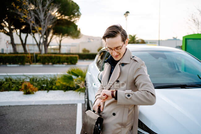 Вид сбоку стильного мужчины-предпринимателя, стоящего возле машины на стоянке и проверяющего время на наручных часах в ожидании встречи — стоковое фото