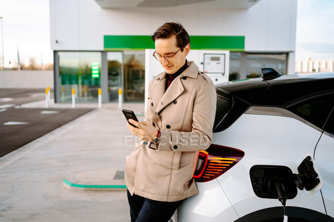 Hombre empresario en traje elegante de pie en la gasolinera y el teléfono móvil de navegación, mientras que de pie cerca del coche con la boquilla de combustible - foto de stock