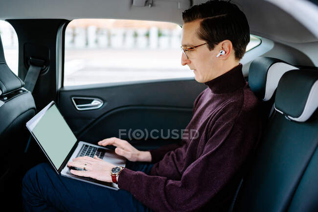 Seitenansicht des geschäftigen männlichen Unternehmers in drahtlosen Kopfhörern, der im Luxusauto auf dem Beifahrersitz sitzt, während er Laptop benutzt und an einem Geschäftsprojekt arbeitet — Stockfoto