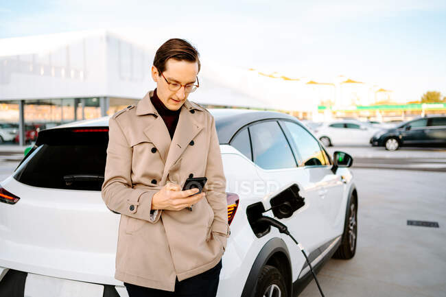 Hombre empresario en traje elegante de pie en la gasolinera y el teléfono móvil de navegación, mientras que de pie cerca del coche con la boquilla de combustible - foto de stock