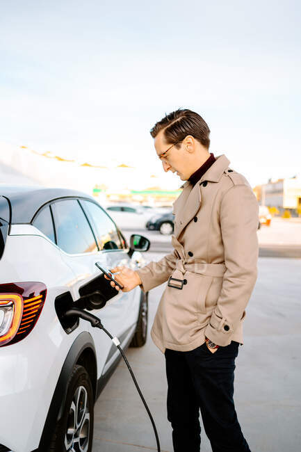 Вид збоку стильного чоловічого підприємця, який переглядає смартфон, стоячи на бензозаправці та дозаправляючи автомобіль — стокове фото