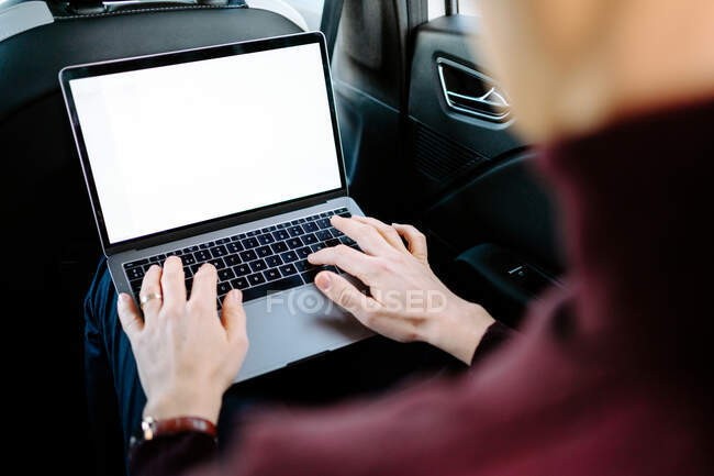 Side view of busy male entrepreneur in wireless earphones sitting in luxury car on passenger seat while using laptop and working on business project — Stock Photo