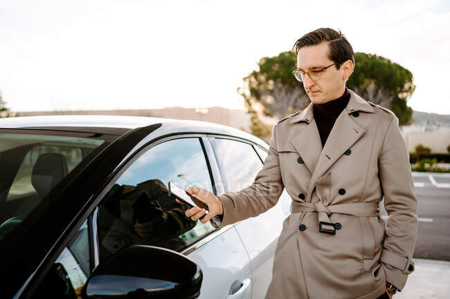 Empreendedor masculino destrancando a porta do automóvel moderno com smartphone enquanto usa o aplicativo de controle remoto — Fotografia de Stock